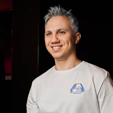 A smiling man with noticeably graying hair wears a white sweatshirt featuring a small cartoon graphic on the chest, illustrating the "more gray hair" update referenced in his day 14 trading challenge recap showing a balance increase from $400 to $459.