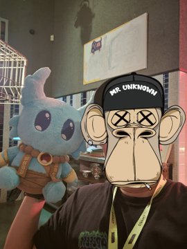 A person wearing a "MR. UNKNOWN" cap poses with a blue Koda plush toy from the Bored Ape Yacht Club NFT collection, standing in front of a monitor displaying a BAYC logo. The image ties to the post by visually representing the "Koda" the author mentions winning in a dream, celebrating the BAYC community vibe.