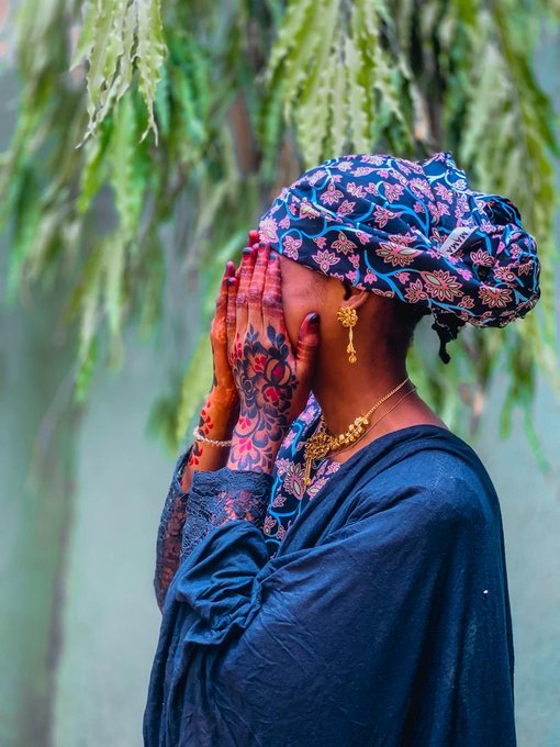 A side profile of a woman wearing a blue patterned headwrap and matching blue garment, with henna-decorated hands covering her face against a leafy green background.