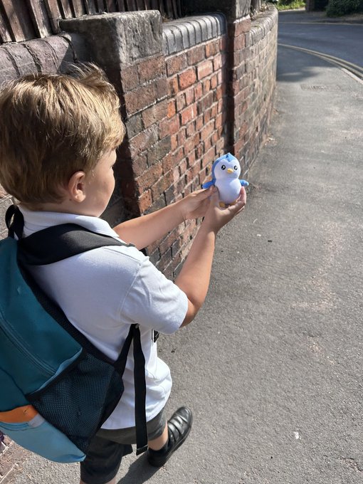 A young boy wearing a backpack holds up a small blue and white penguin plush toy while walking alongside a brick wall.