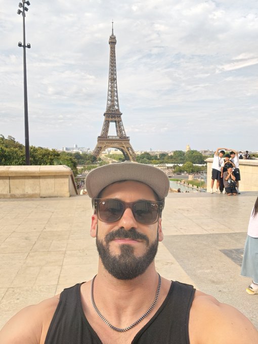 A bearded man wearing sunglasses, a gray cap, and a black tank top takes a selfie with the Eiffel Tower in the background in Paris.