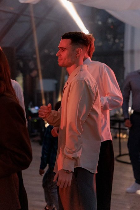 A man in a light-colored button-up shirt standing in profile under warm red lighting at what appears to be an indoor event or venue.