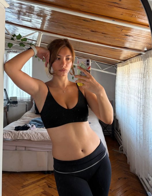 A young woman takes a mirror selfie in a bedroom wearing a black sports bra and dark gray leggings, holding her phone up to capture the photo.
