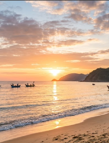 A serene beach sunset scene with golden sunlight reflecting on calm ocean waters, distant boats, and silhouetted mountains under a partly cloudy sky.