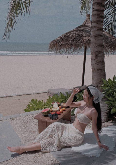A woman in a white lace outfit sits on a towel on a sandy beach next to a small table with fruit, with the ocean and a palm-thatched umbrella in the background.