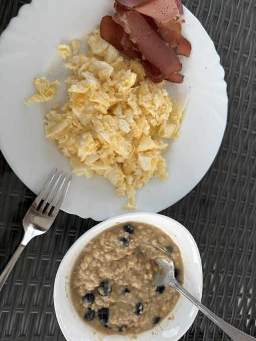A top-down photograph of a breakfast plate containing scrambled eggs and bacon, accompanied by a side bowl of oatmeal topped with blueberries and a fork resting on the edge.