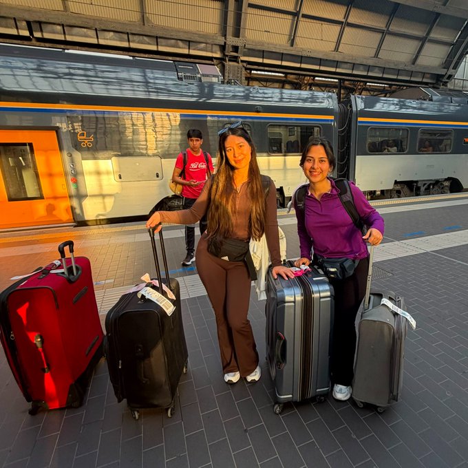 The image shows two women standing next to their luggage at a train station, with an orange train visible behind them. They are posing for a photo while holding their suitcases. The text "Milan to Lugano" can be seen near the bottom of the image, which suggests that they might be traveling from Milan to another city in Italy and then to a neighboring country or region.