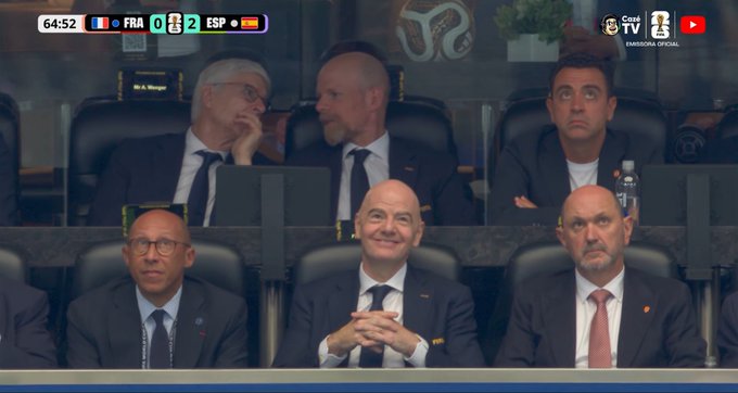 Gianni Infantino, FIFA president, is seen smiling in a VIP box during the France vs Spain match, which ended with a 0-2 scoreline as shown on the scoreboard at the 64:52 mark. The image relates to the "#FRAXESP" post by capturing Infantino's reaction during the game, with his smile hinting at the reason behind the post's tone.