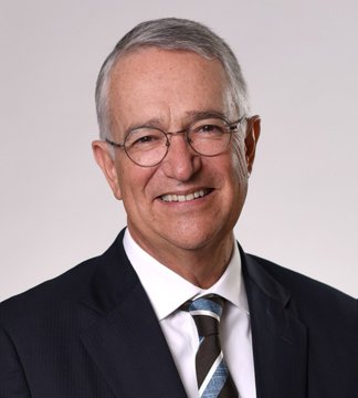 A professional headshot portrait of a smiling older man with gray hair, wearing glasses, a dark suit jacket, white shirt, and a blue patterned tie against a plain white background.