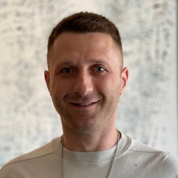 The image features a man wearing a white shirt who has a chain around his neck. He is smiling at the camera, giving off an impression of happiness or positivity. The background behind him appears to be a wall with some writing on it. There are also several other people in the image, but they are not the main focus and do not seem to have any direct connection to the man wearing the chain around his neck.