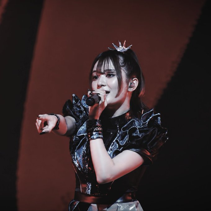 A Japanese pop performer (resembling BABYMETAL's Moona) singing on stage wearing a black ruffled outfit and a small crown.