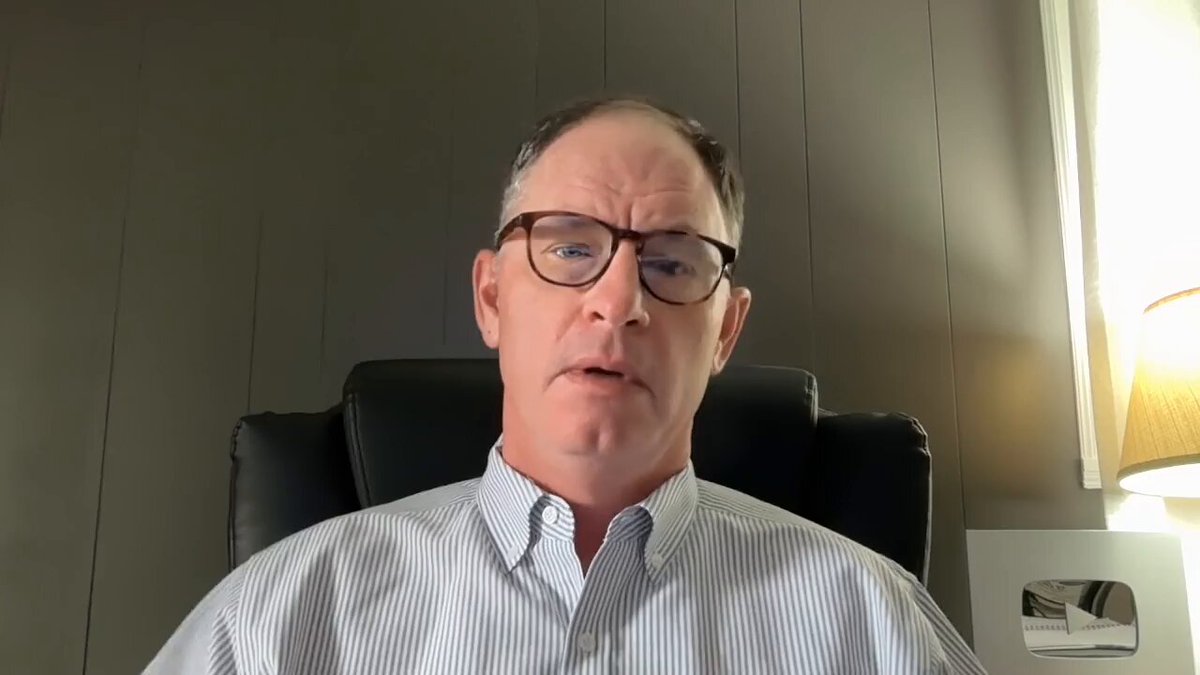 The image shows a man wearing glasses sitting in an office chair, looking at the camera with a serious expression. He appears to be engaged in a video call or a live stream, as indicated by the presence of a laptop computer placed on his lap. The background features a wall and a window, suggesting that he is likely working from home or in a professional setting.