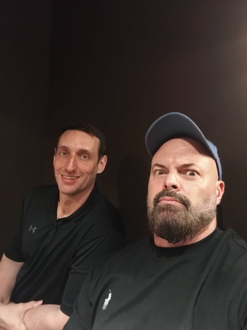 A selfie-style photo of two men; the one on the right wears a blue baseball cap and a black polo with a pony logo and has a thick beard, while the man on the left smiles and wears a black polo with an Under Armour logo.