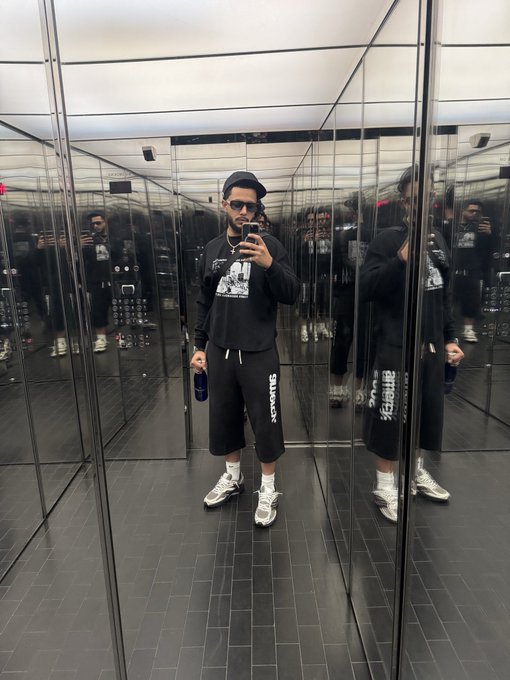 A person takes a mirror selfie in a mirrored room, wearing a black graphic hoodie, black shorts with white text, a bucket hat, sunglasses, and chunky sneakers.