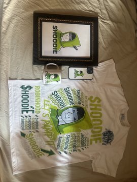 A collection of $HOODIE branded merchandise including a framed picture, t-shirts with green character artwork, and stickers laid out on a bed.