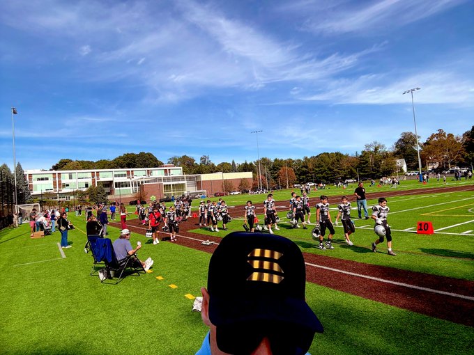 An outdoor football field scene with players in uniform running drills, viewed from behind a spectator wearing a dark cap, with a school building in the background under a blue sky.