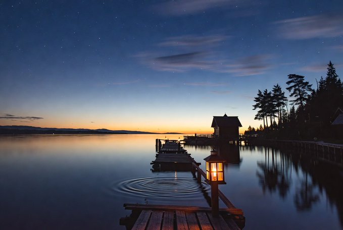 The image features a dock at night, with a small wooden cabin sitting next to it. The cabin has a lantern hanging from its roof, providing light for the surrounding area. In the background, there are trees that add to the serene atmosphere of the scene. The caption provided seems unrelated and does not provide any additional information about the image or its context.