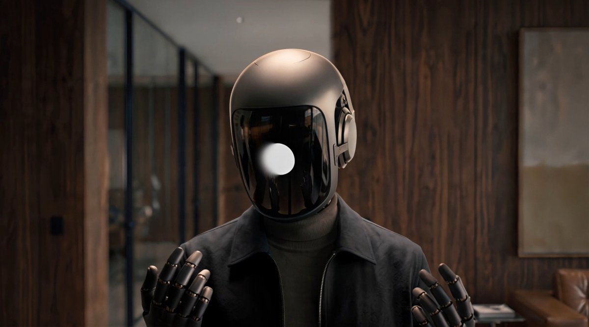 A figure wearing a metallic, reflective helmet with a dark visor, a dark jacket, and robotic gloves, standing in a wood-paneled room.