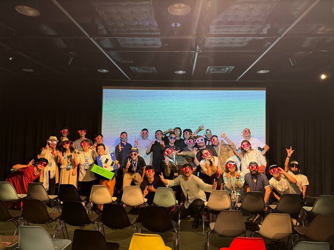 A large group photo of approximately 30 people wearing colorful eye masks and casual clothing posing cheerfully on a stage with a large LED screen behind them.