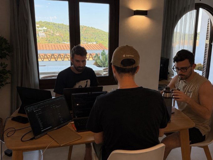The image shows a group of three men sitting at a table, each working on their laptop computers. They are all wearing baseball caps and appear to be focused on their work. The room has large windows that provide natural light, creating a comfortable atmosphere for productivity. One man is holding a cup in his hand, suggesting they might be enjoying some refreshments while working or discussing projects.