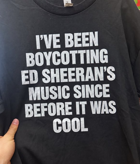 A black t-shirt with white text reading 'I'VE BEEN BOYCOTTING ED SHEERAN'S MUSIC SINCE BEFORE IT WAS COOL' being held up by someone's hand.