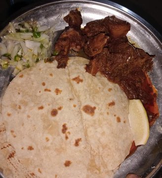 A close-up of a metal plate holding what appears to be tacos or a meat dish with flatbread tortillas, a side of onions and cilantro, and a lemon wedge.