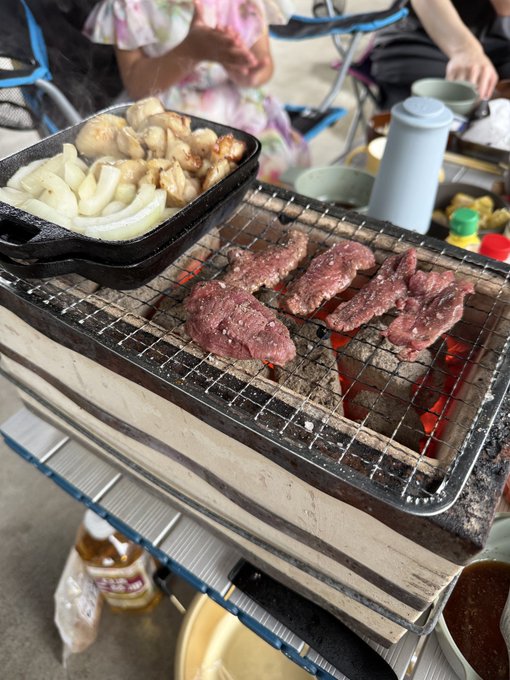 Strips of raw beef and onions grilling on a charcoal grill at an outdoor cooking setup, with a bottle of sauce and other items nearby.