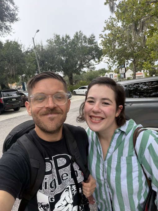 A selfie of a smiling couple—a man with glasses and a mustache wearing a dark t-shirt, and a woman in a green and white striped shirt—standing outdoors with trees and parked cars behind them.