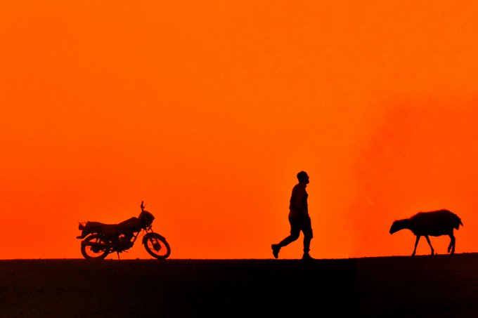 The image features a man walking across a road, with his shadow cast on the ground behind him. A motorcycle is parked nearby, and another person can be seen further back in the scene. The background of the photo has an orange hue, suggesting that the sun is setting or it's early morning. The post contains text that reads "I have just collected Sunset by @Abbas_rahmani63 on @objktcom". This implies that the image was taken during sunset and features a man walking across the road while his shadow is cast behind him.