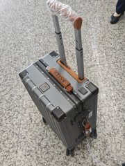 A dark gray hardshell suitcase stands upright on a gravel surface, featuring extended telescoping handles wrapped in protective plastic.