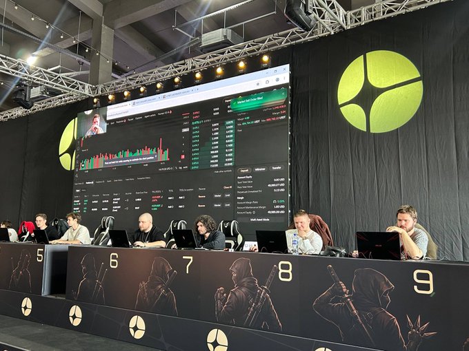 Players compete at a gaming tournament setup with numbered stations 5 through 9, facing a large screen displaying market data and gameplay under a 'DOTA 2' logo.