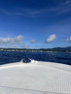View from the bow of a boat on calm blue water with mountains and a coastal town in the distance under a partly cloudy sky.