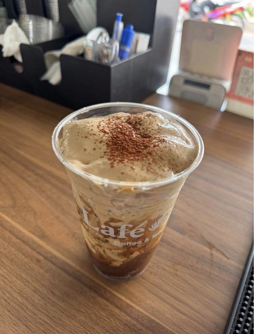 An iced coffee or blended beverage in a clear plastic cup with 'cafe Coffee &' printed on it, topped with foam and a sprinkle of cocoa powder, sitting on a wooden café table.