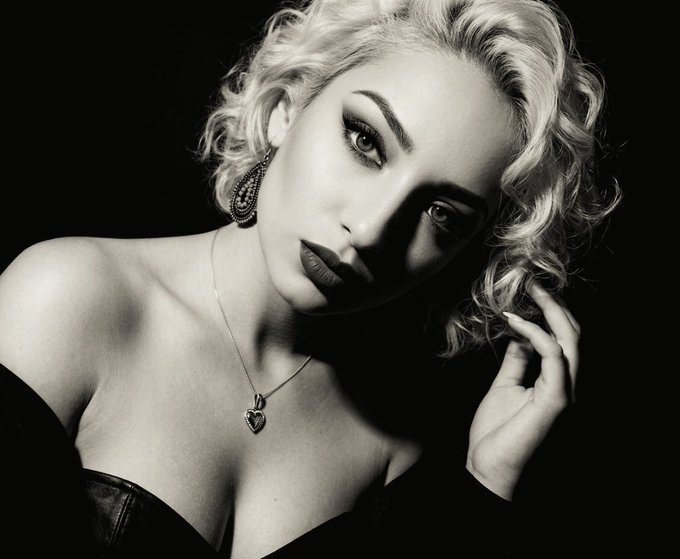 A black and white glamour portrait of a woman with curly blonde hair, dramatic makeup, ornate earrings, and a heart pendant necklace against a dark background.