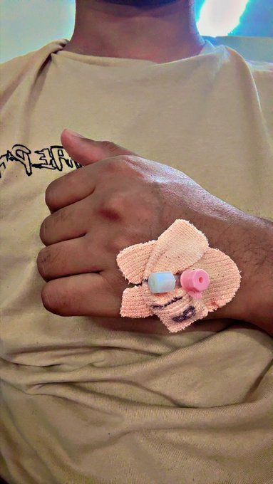 A close-up photo shows a person's hand with a small medical IV line and a pink bow bandage attached, giving a thumbs-up gesture.