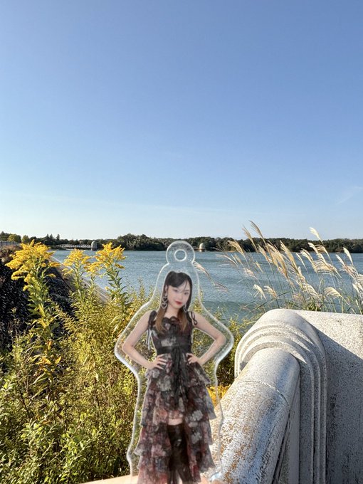 An acrylic standee of a K-pop idol posed outdoors with hands on hips, set against a scenic background featuring a body of water, tall grass, and a clear blue sky.