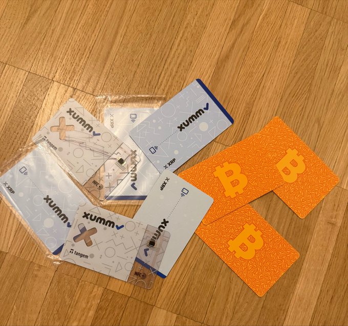Two Xaman (formerly Xumm) Tangem hardware wallets and two orange Bitcoin-themed cards lie scattered on a wooden floor, illustrating the user's collection of physical crypto storage devices including their preferred XRPL wallet alongside alternative options.