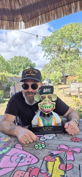 A bearded man in sunglasses and a 'WORLD FAMOUS' cap posing with a quirky tiki-style statue at an outdoor bar.