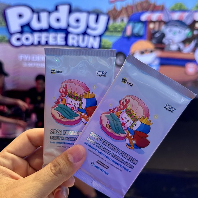 A hand holding two Pudgy Penguins trading card packs in front of a 'Pudgy Coffee Run' promotional banner, with text reading '2026 KAKAO X PUDGY TRADING CARDS'.