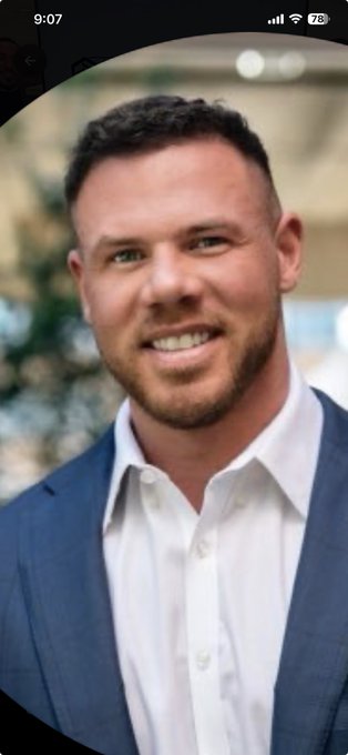 A professional headshot of a smiling man with short brown hair and a beard, wearing a white dress shirt and blue blazer.