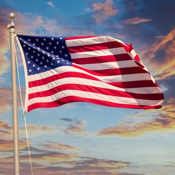 The American flag waving on a flagpole against a dramatic sunset sky with orange and blue clouds.