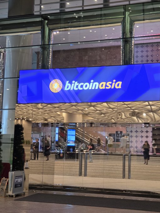 A brightly lit venue entrance displays a large blue banner reading "bitcoinasia," indicating the Bitcoin Asia conference is taking place in Hong Kong, where attendees gather to share developments like the Power Law model within the crypto community.