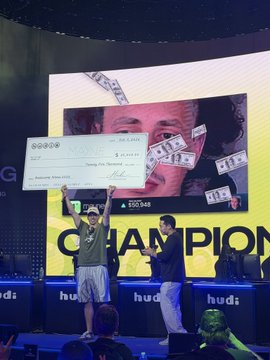 A man holding up an oversized novelty check on stage in front of a screen displaying a face and falling money graphics, with 'CHAMPION' and 'hudl' visible below.