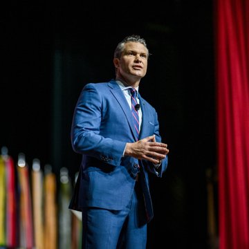 Defense Secretary Pete Hegseth, shown speaking on stage in a blue suit, is at the center of reports that his annual financial disclosure revealed Bitcoin holdings in a Coinbase wallet valued between $16,000 and $65,000.