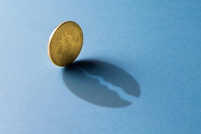 A photograph of a gold Bitcoin coin standing upright on a blue surface, casting a distinct shadow.