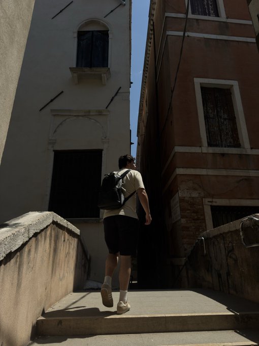 A person wearing a white t-shirt, black shorts, and a backpack walking up concrete stairs between two buildings with warm terracotta-colored walls under bright daylight.