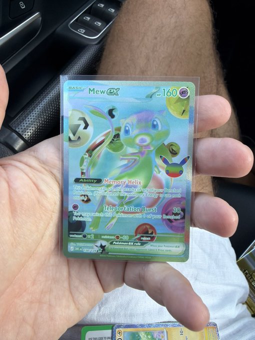 A hand holds a holographic Mew ex Pokémon trading card (#160) inside a vehicle.