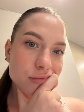 A close-up selfie of a young woman with blue eyes and dark hair pulled back, resting her chin on her hand.