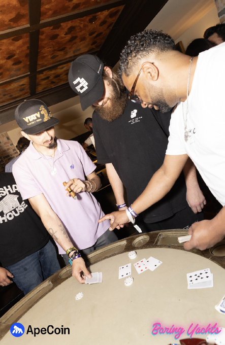 A promotional photo from an 'ApeCoin' sponsored 'Boring Yachts' event showing several men gathered around a casino-style card table, with one bearded man in a black cap dealing cards while others watch closely.