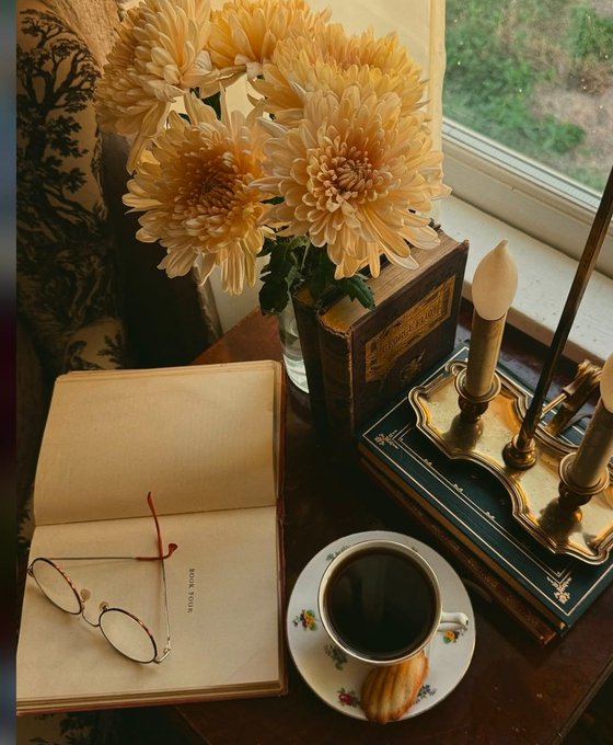 An aesthetic still life featuring an open book with wire-rimmed glasses resting on it, a teacup of dark tea, peach-colored chrysanthemums in a vase, vintage books, and brass candlesticks near a window.